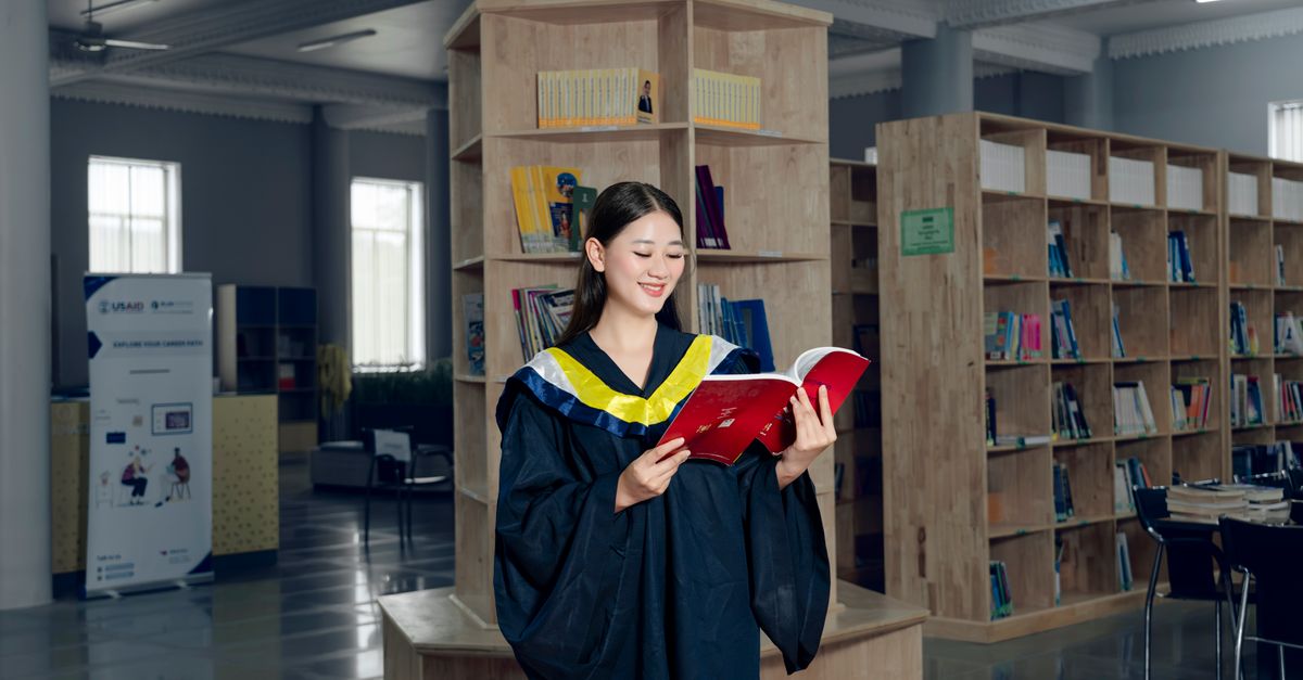 Woman in graduation gown reading a book in university library, symbolizing education and knowledge.